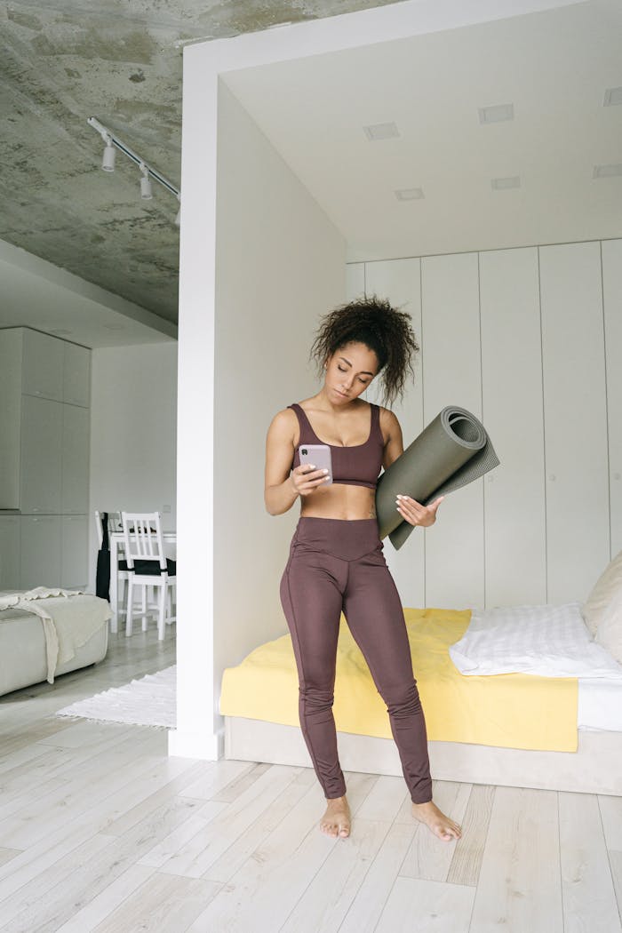 gallery-3 Woman in activewear standing with yoga mat and smartphone in a modern bedroom setting.